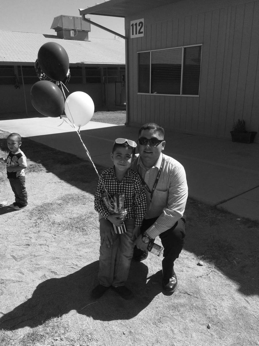 An adult kneeling beside a child holding balloons outside a school building.