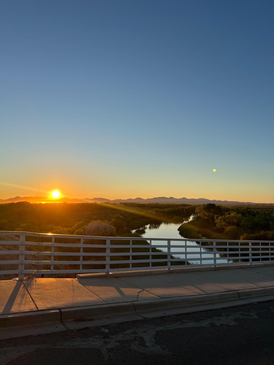 Sun setting over a river beside a bridge.